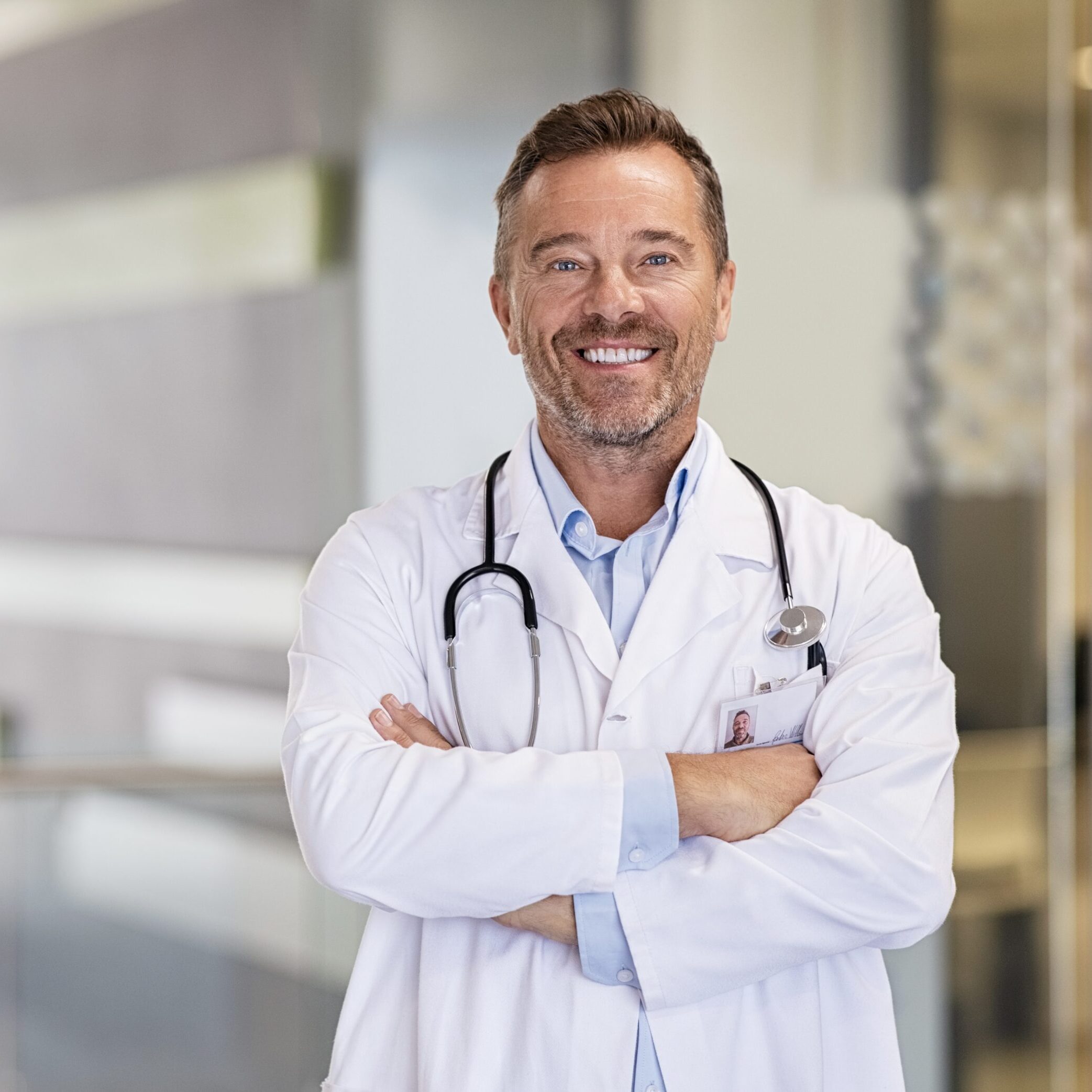 Portrait of happy mature doctor with folded arms standing at hospital hallway. Confident male doctor in a labcoat and stethoscope looking at camera with satisfaction. Smiling confident general practitioner at private health clinic.